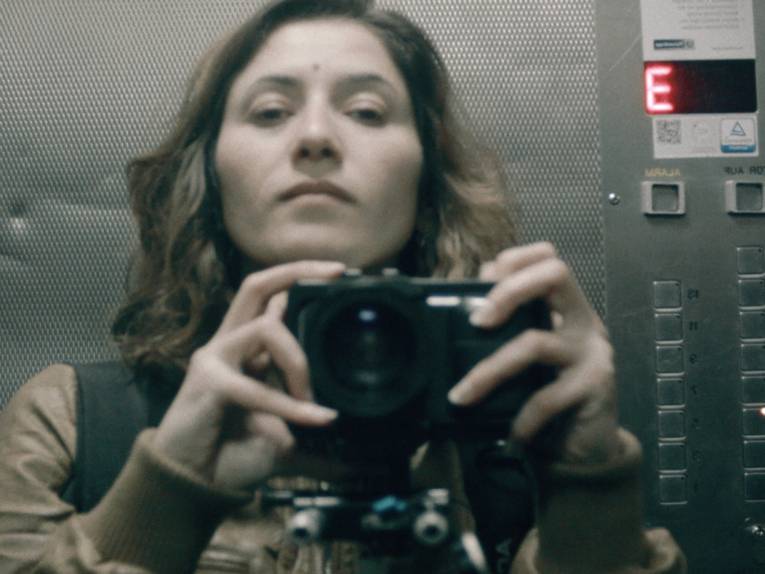 Eine Frau in einem Fahrstuhl. Sie hält mit beiden Händen einen Fotoapparat und schaut in die Kamera